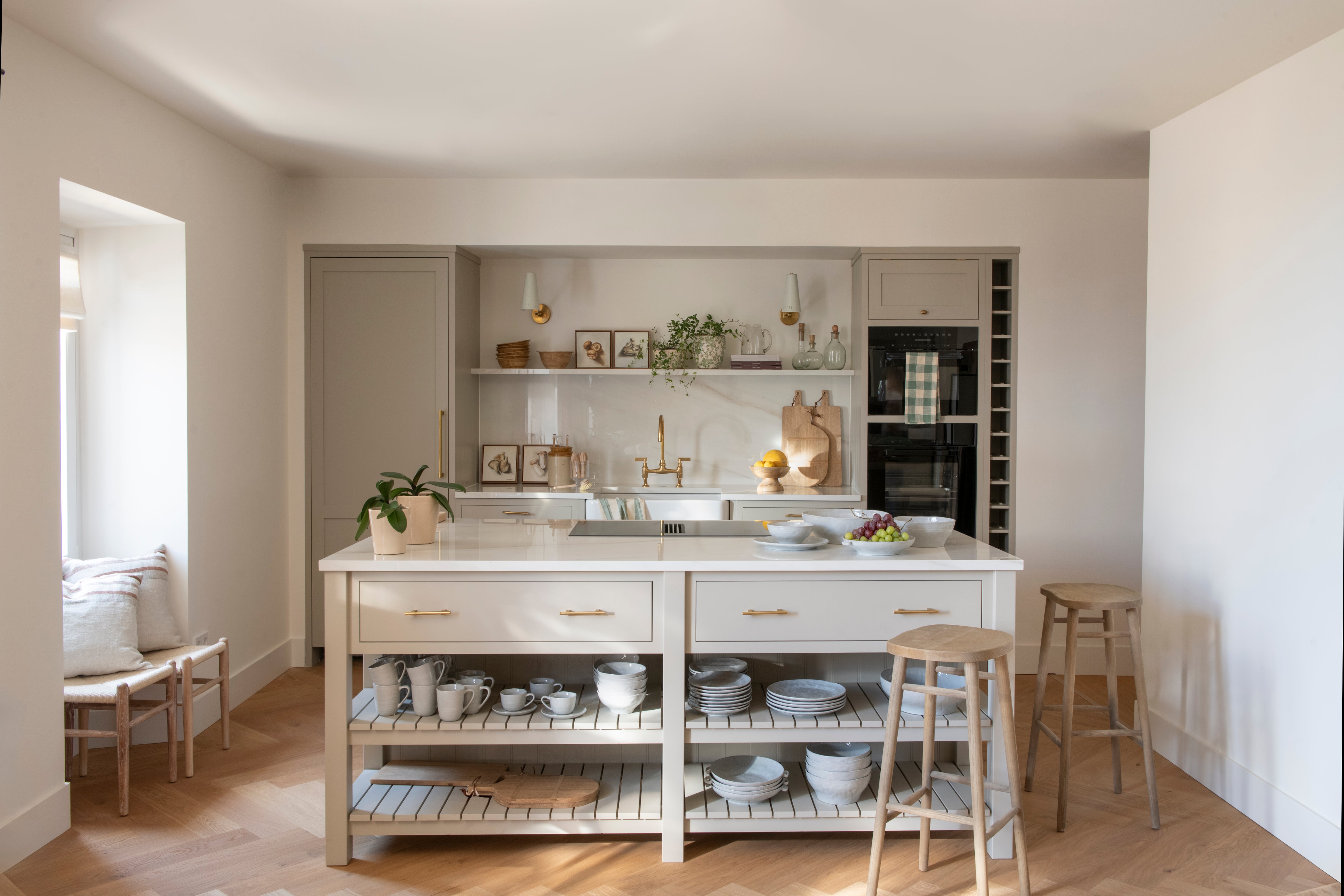 Wide view of a light and neutral kitchen with a central island, open shelving, and a polished brass bridge mixer on the back counter.