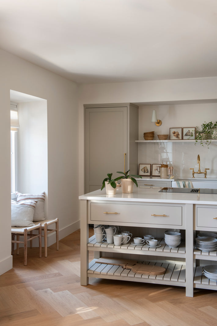 Styled kitchen scene showing the polished brass bridge mixer framed by natural wood accents, soft neutral cabinetry, and decorative objects.