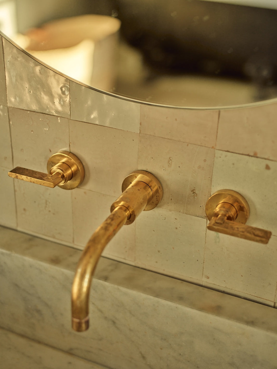 A three hole wall mounted basin tap in unlacquered polished brass is positioned above a marble sink, against white zellige tiles. 