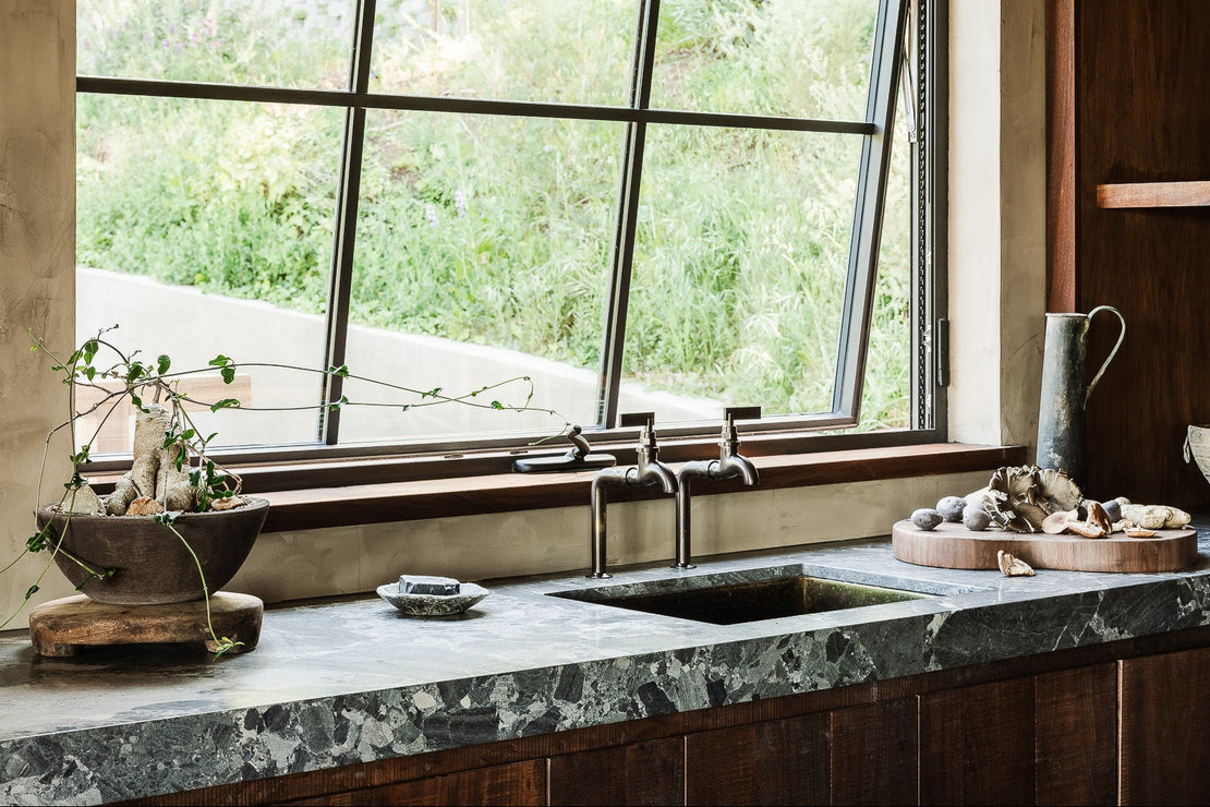 Modern kitchen with marble countertop, wooden cabinets, and a large window. Two brass bib taps by Studio ORE.