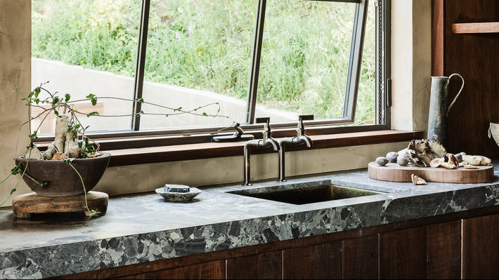Modern kitchen with marble countertop, wooden cabinets, and a large window. Two brass bib taps by Studio ORE.