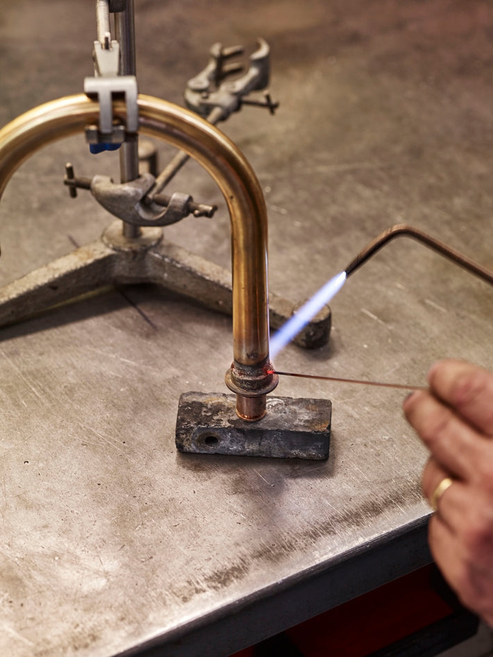 A curved spout being hand-finished in Studio Ore's workshop. The spout is from Chapter Two, and the craftsman is using a mini blow torch to sharpen the details. 