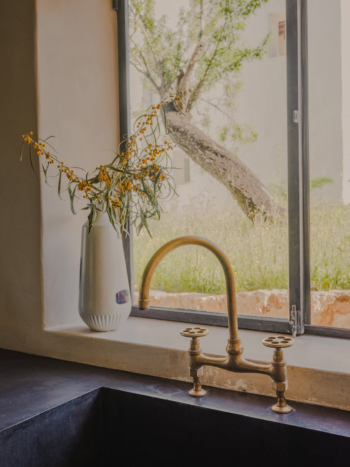 In a modern Ibizan home, Studio Ore's Kitchen Bridge Mixer Tap in unlacquered polished brass stands over a wide basin.