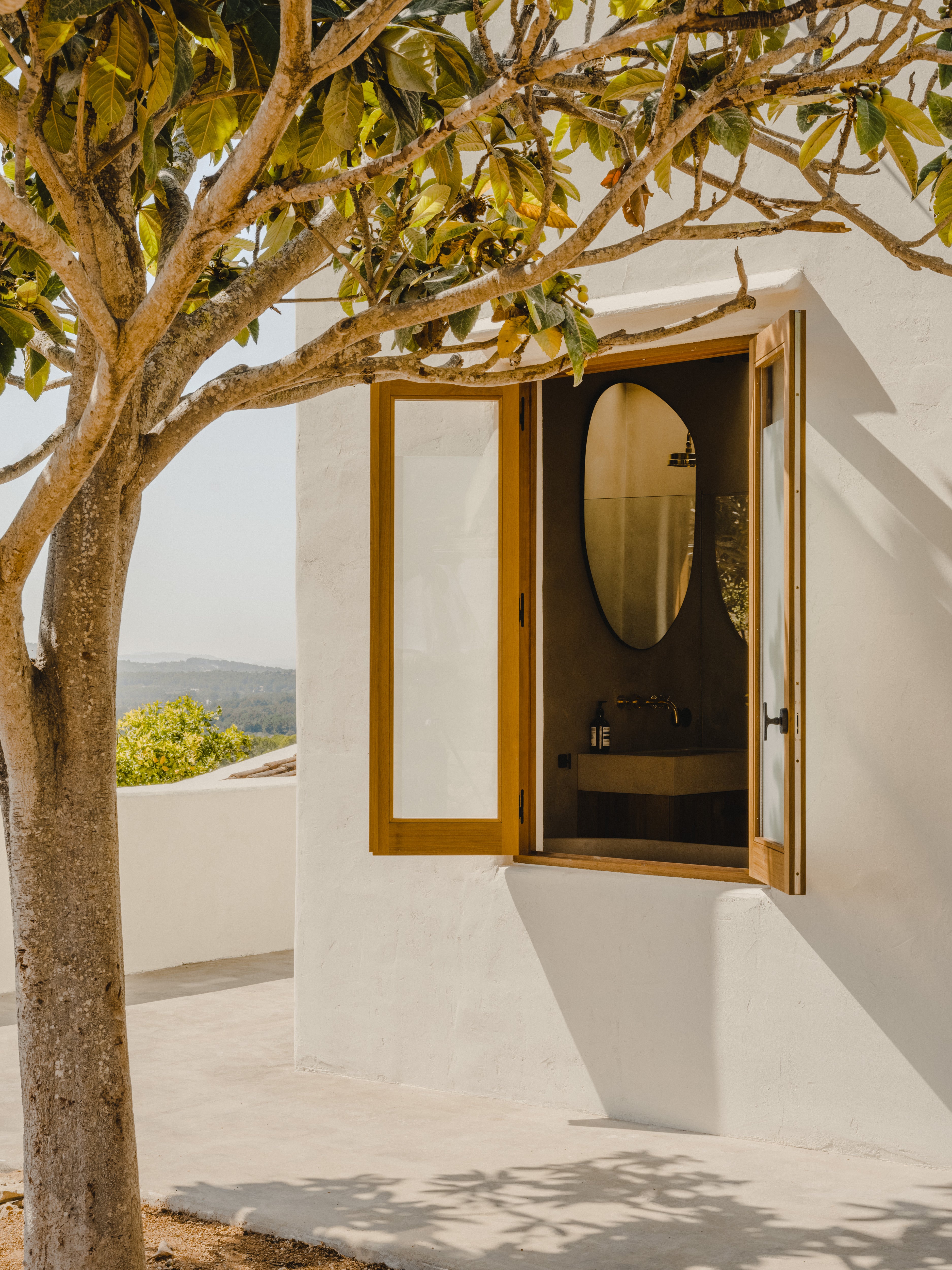 Exterior view into bathroom at Ibiza Campo Casa, showing Studio Ore brass wall mounted basin tap in Unlacquered Brass, framed by whitewashed walls and timber windows.