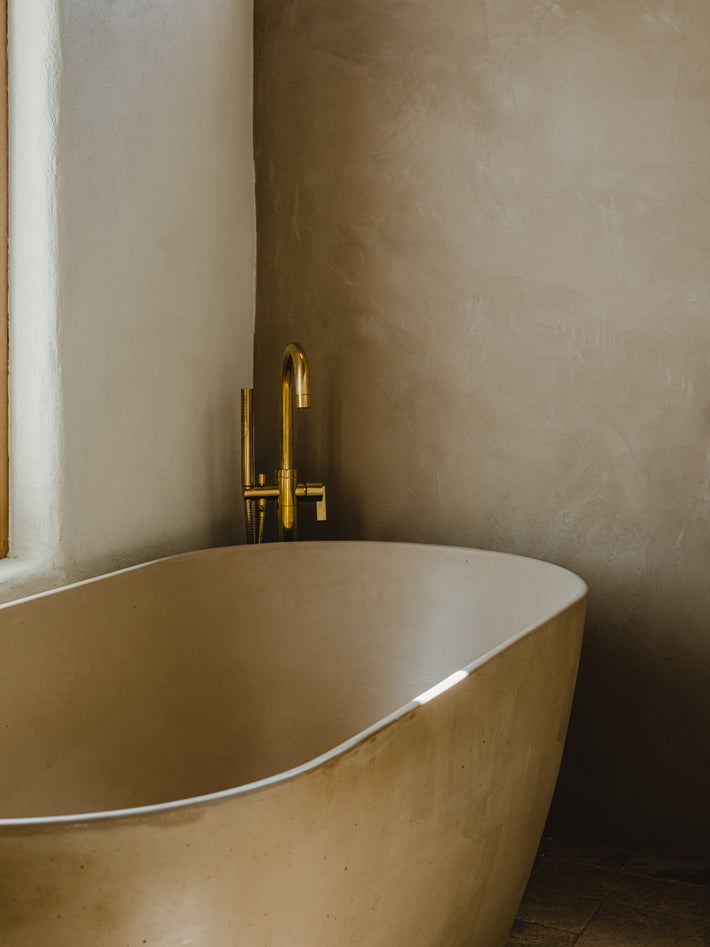 Freestanding stone bath with Studio Ore floor mounted bath mixer in Unlacquered Brass at Ibiza Campo Casa, with plaster walls and natural light.