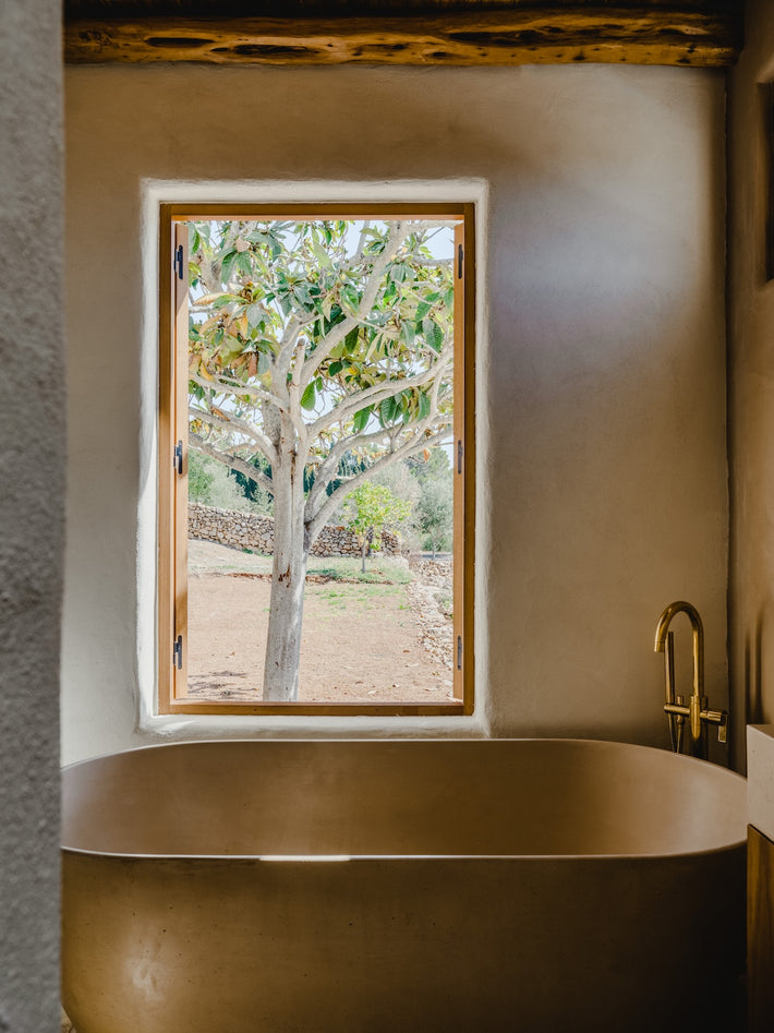In the same bathroom, a freestanding concrete basin sits below a large window looking out to the Ibiza countryside. Studio Ore's bath mixer stands over the bath, also in polished brass. 