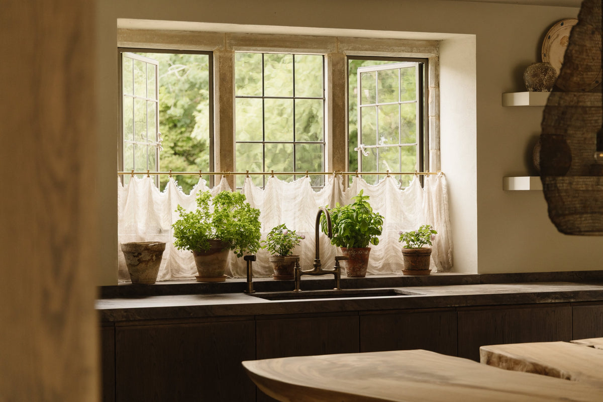 Studio Ore's deck-mounted kitchen bridge mixer and kitchen hand rinse in unlacquered tumbled bronze. The fittings stand in a rustic kitchen. 
