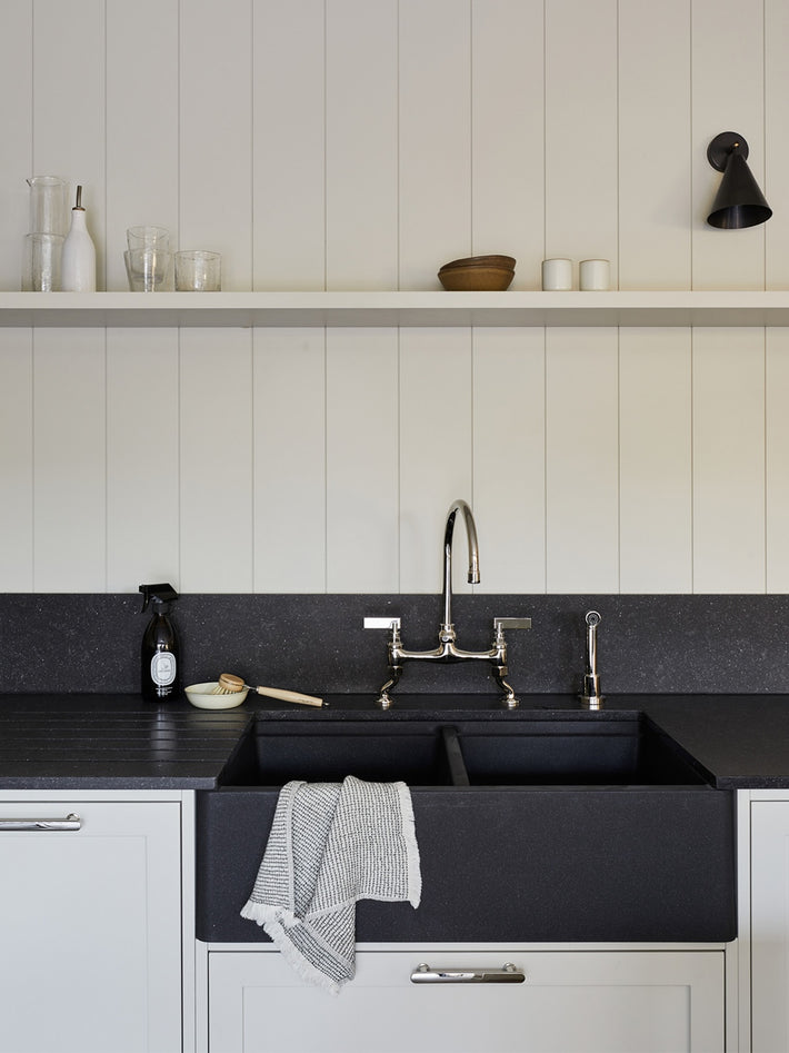 Studio Ore fittings are found in this project by Studio Skey. in Polished nickel, the kitchen bridge mixer stands on a black marble worktop and double basin. Next to it, the hand rinse. The kitchen is monochromatic with white panelled walls and units. 