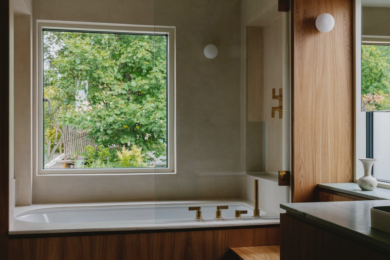 Studio Ore fittings can be seen in a modern bathroom designed by Studio Hagen Hall. There's bath controls, with an overhead shower above and wall mounted thermostat. A large, square window is above the bath which looks out to the garden. 