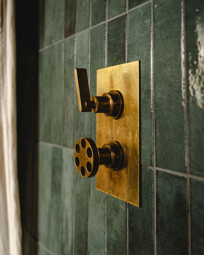 Close-up of brass shower thermostat controls mounted on green tiled wall.