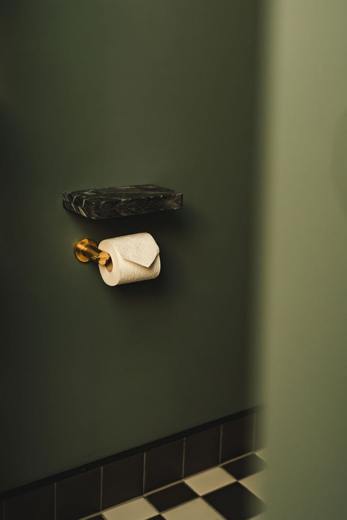 Brass toilet roll holder with marble shelf above, set against green painted wall.