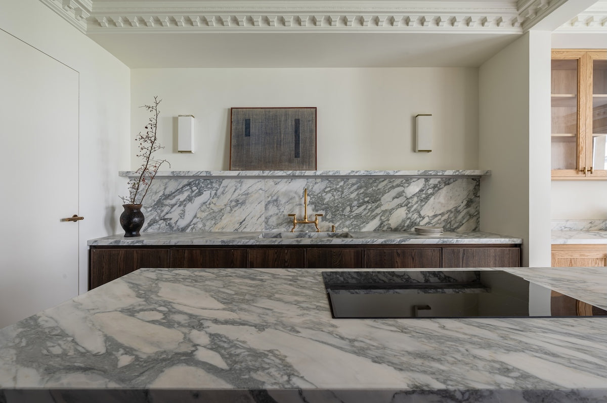 Studio Ore's kitchen bridge mixer tap in unlacquered polished brass stands in a  kitchen with marble surfaces.